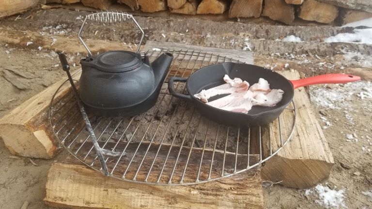 Bacon cooking in a cast iron skillet over hot coals. Water boiling in a black cast iron pot for cowboy campfire cooking equipment. 