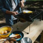 From above of crop anonymous male cooker frying eggs on skillet using metal stove on table in forest