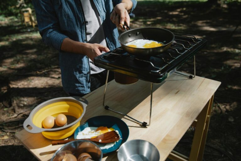 From above of crop anonymous male cooker frying eggs on skillet using metal stove on table in forest