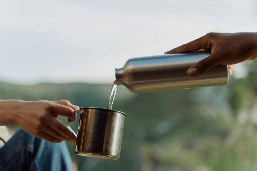 Close-up of hands pouring water from a metal bottle into a cup in an outdoor setting.